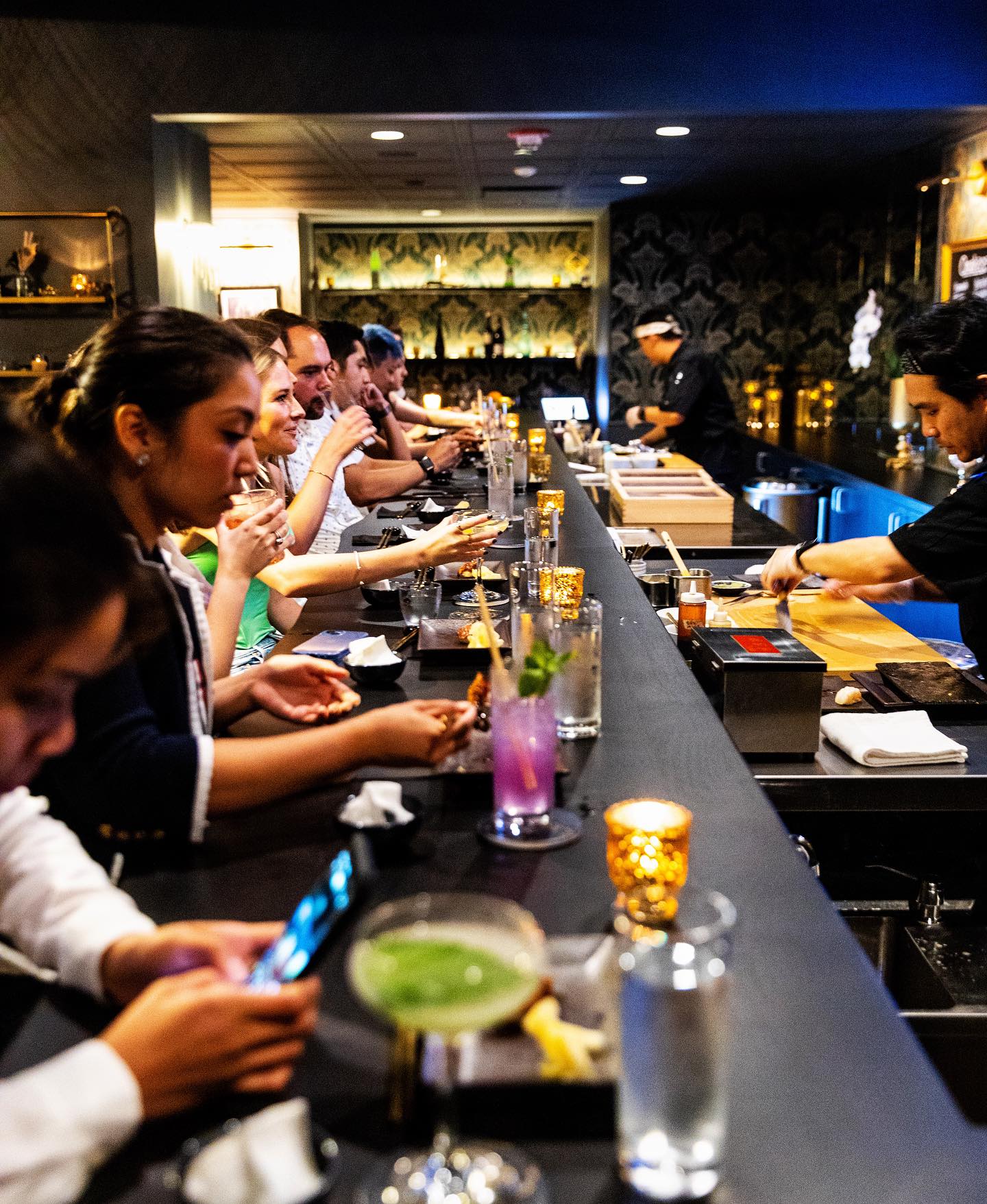 Row of people at left dine on sushi and people at right preparing food
