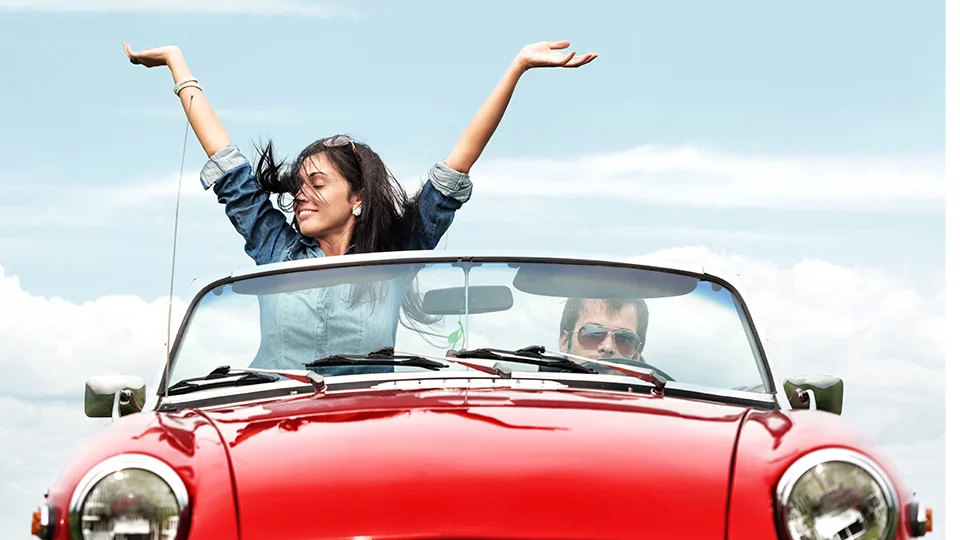 Woman with outstretched arms in red convertible, man at the wheel