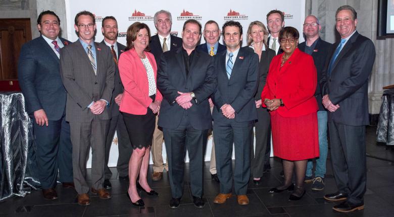 BID board members dressed in fancy business attire stand in front of a step and repeat board, smiling at the camera. 
