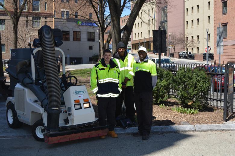 Three employees in reflective jackets stand in front of a park, facing the camera with a smile. A street sweeper on their left. 