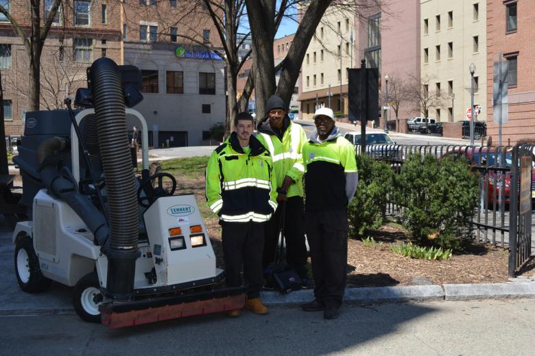 Three employees in reflective jackets stand in front of a park, facing the camera with a smile. A street sweeper on their left.