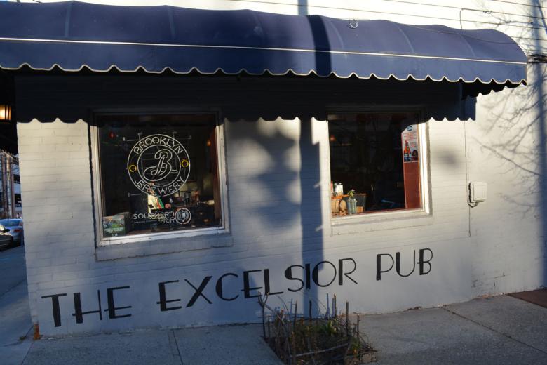 Exterior shot of the Excelsior Pub building, blue awning over the entrance with the logo painted on the building. 