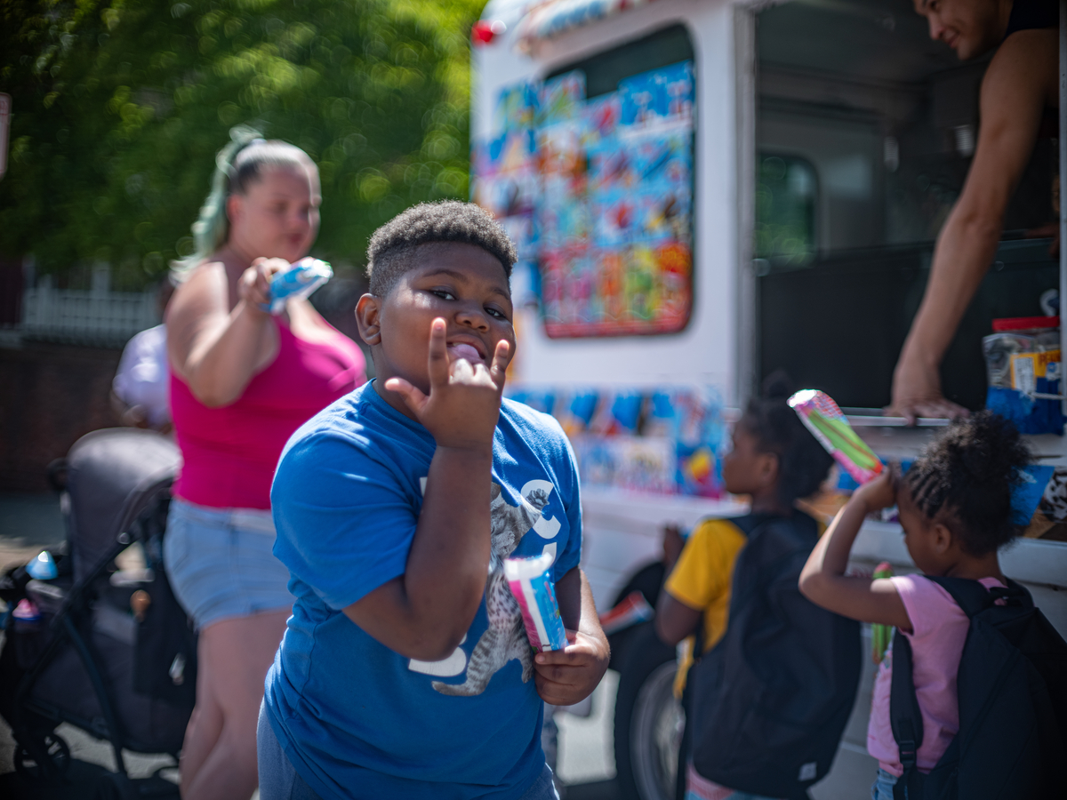 Boy looking at camera and holding up I Love You sign, and in background people stand at ice cream truck.