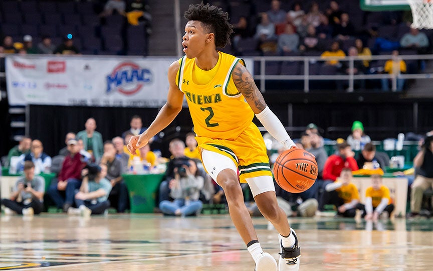 Siena Saints College basketball player in yellow and green uniform dribbling a ball.