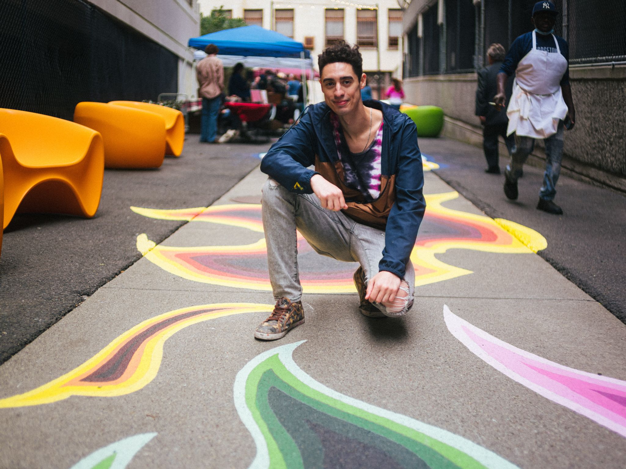 Man crouches down on sidewalk mural with people and outdoor furniture in background