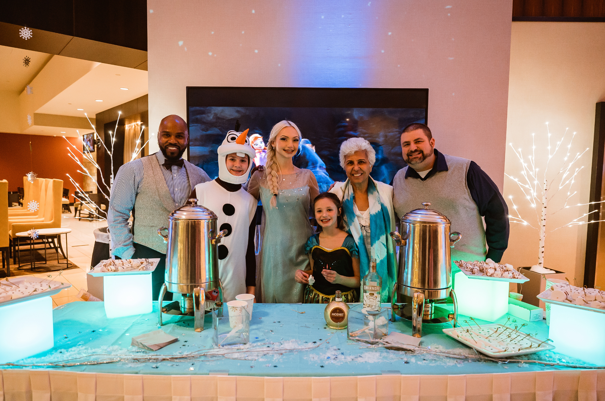 Six hotel staff members dressed up as characters from the movie Frozen, posing for a picture behind a hot chocolate stand in their lobby.