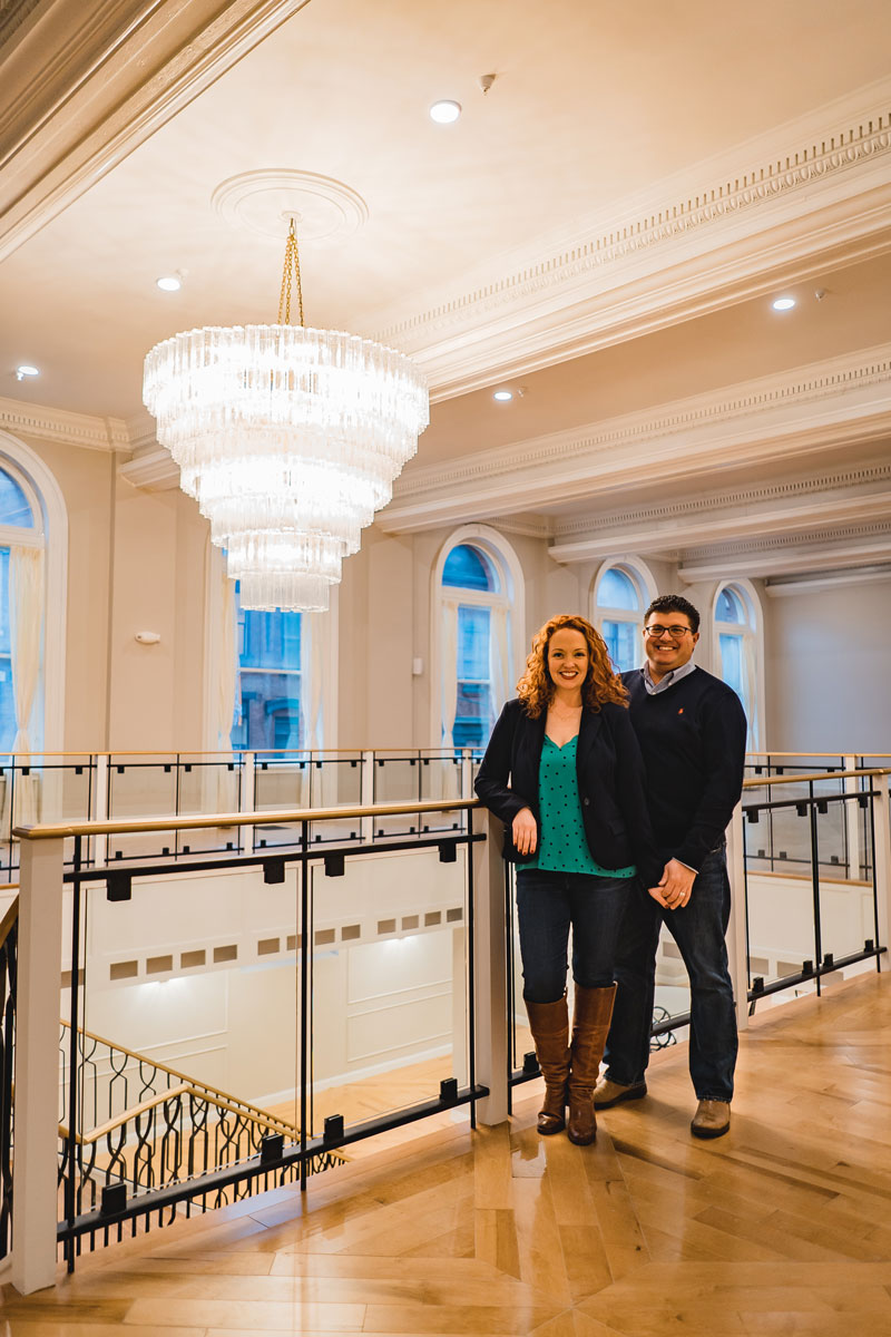 Couple standing on an upper floor, leaning against a railing. A large, elaborate chandelier commands the upper left section of the photo. 