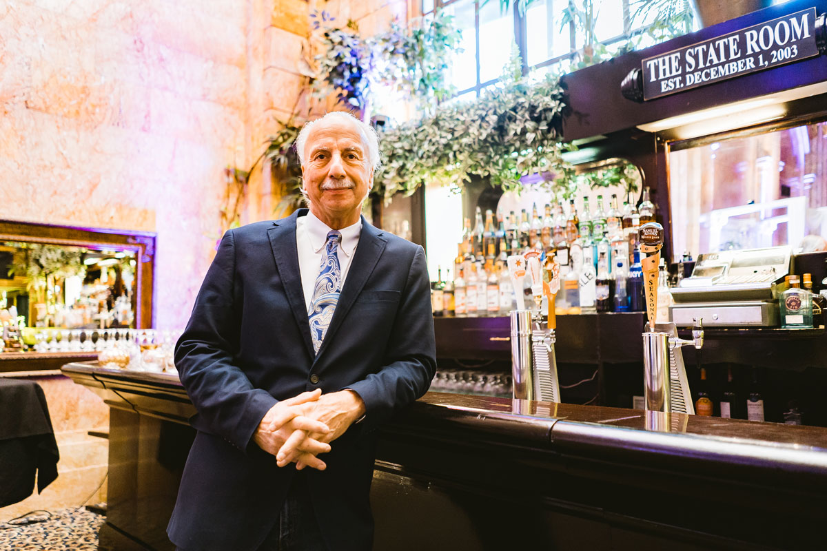 Man in a suit leans agains an old style bar top, The State Room sign showing behind him. 