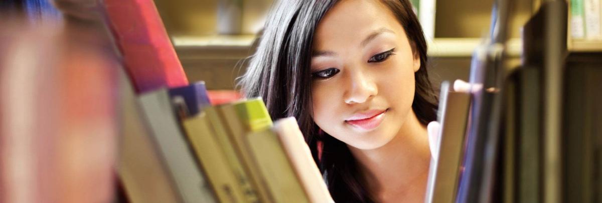 College-age woman looking through books on bookshelf