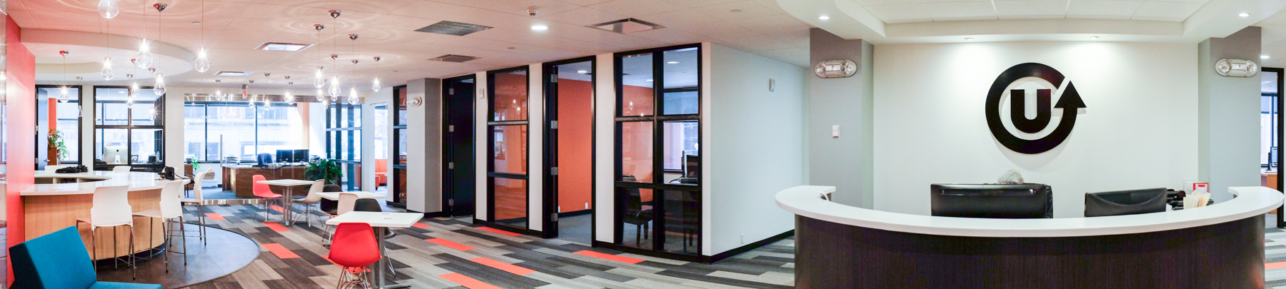 Interior panorama of the Upside Collective offices. A grey and orange blocked carpet is a prominent feature, along with a large decal of their logo on a white wall in the foreground. 