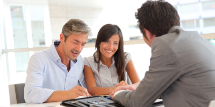 A couple sits at a desk with a man in a suit smiling while signing paperwork. 