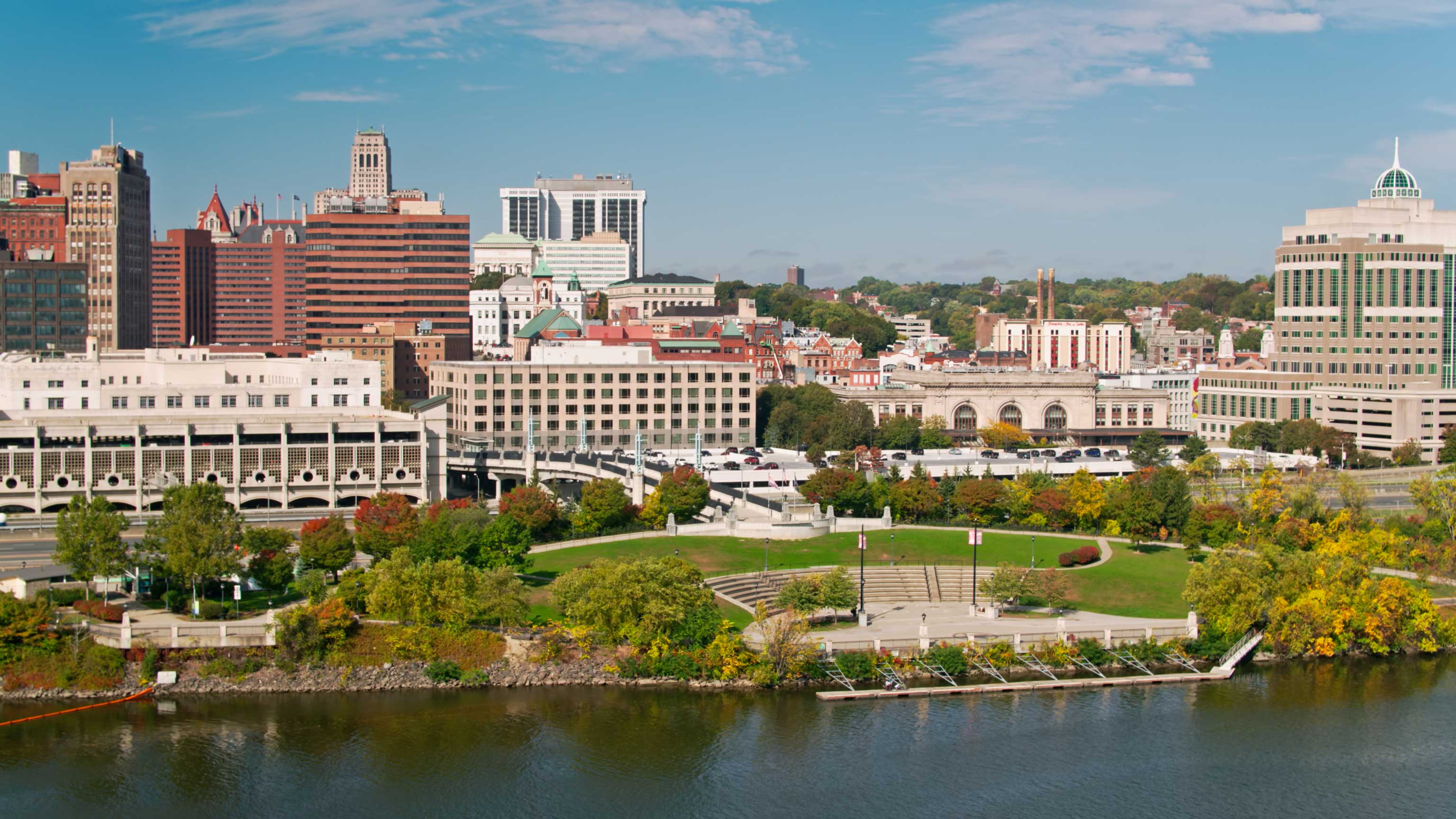 Aerial view of the Corning Preserve and buildings in Downtown Albany with the Hudson River in the foreground. 