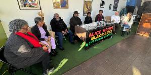 Eight people sit in a line inside building, with the fourth, fifth, and sixth person from left sitting behind a table. On the table hangs a banner reading "Alice Moore Black Arts & Cultural Center".