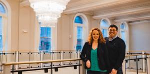 Couple standing on an upper floor, leaning against a railing. A large, elaborate chandelier commands the upper left section of the photo. 