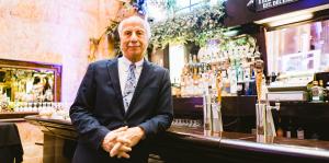 Man in a suit leans agains an old style bar top, The State Room sign showing behind him. 