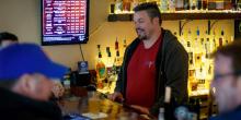 Man stands behind bar facing left, with customers in foreground. 