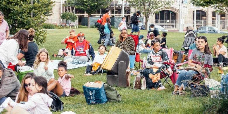 People sit on a lawn on blankets and chairs, with building in background