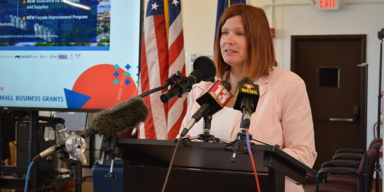 Woman stands at podium speaking into microphones in front of her 