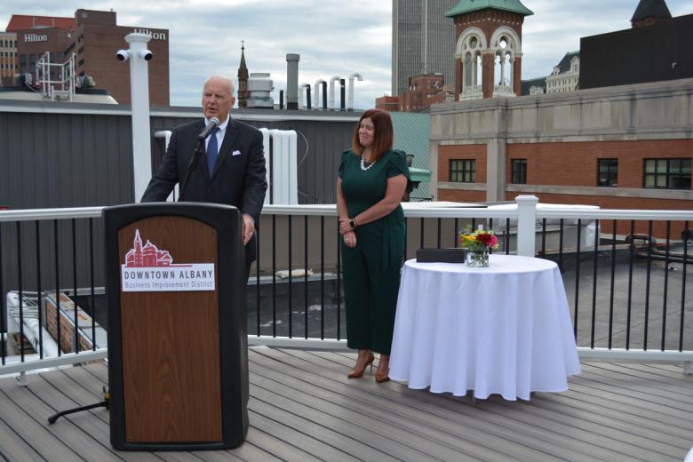 Two people on a rooftop, one standing behind a podium addressing a crowd out of frame, the other standing behind next to a fancy white table