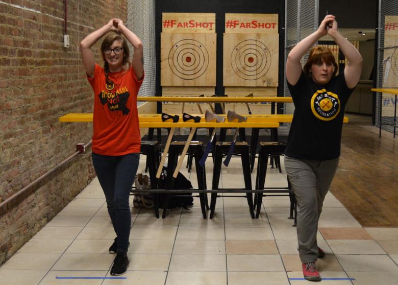 Two people with axes held above their head line up to throw at their targets