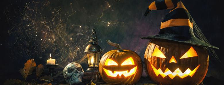 Stock photo of spooky halloween jack-o-lanters wearing witch hats, a skull and candles on the left side of the image and the background is dark with spiderwebs 