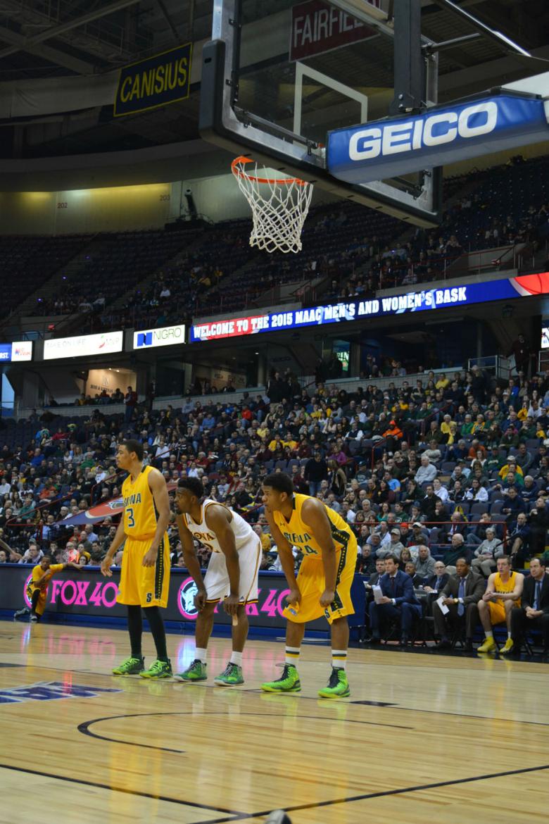 Three basketball players on the court in the middle of a game, the crowd filling the stands behind them. 