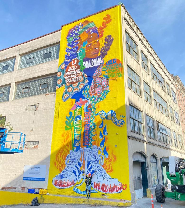 Large mural adorns the side of a building. The illustration is over a bright yellow background and features a mix of portraits and typography.
