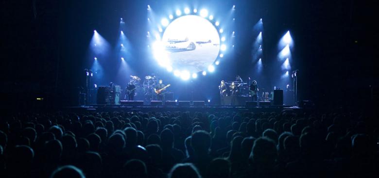 View of a concert stage from the back of the crowd, bright blue lights illuminating the stage and performers. 