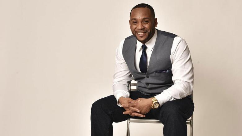 Headshot of Angelo Maddox sitting on a chair in front of a neutral colored background 